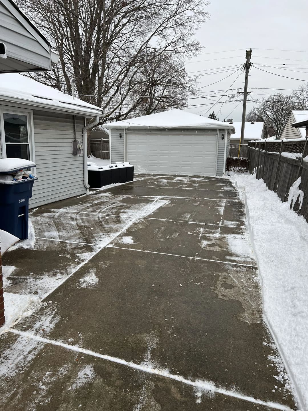 Snow-covered driveway with a residential garage and refuse bin, surrounded by bare trees.