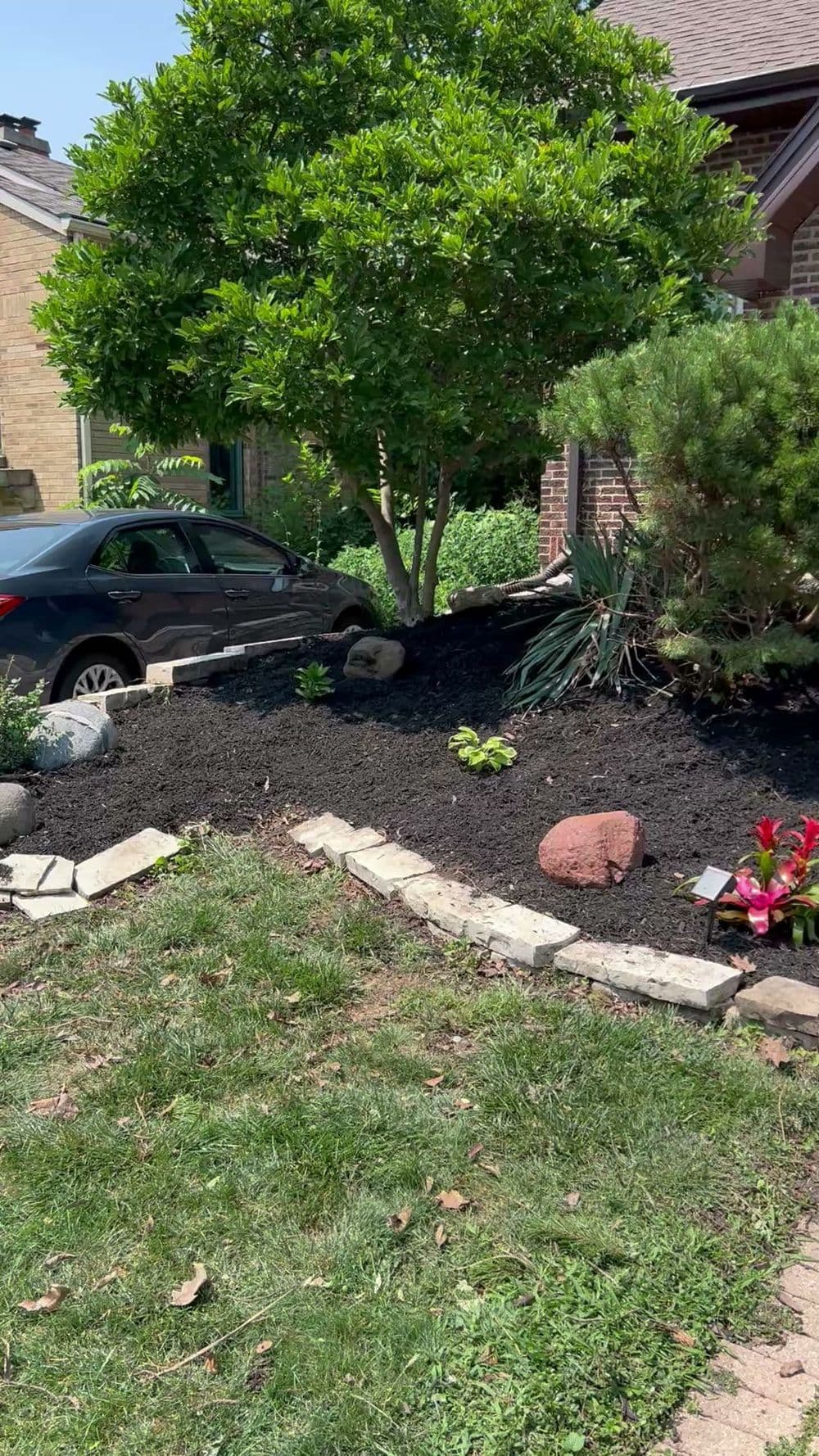 Lush garden landscape featuring plants, mulch, rocks, and a parked car beside a home.